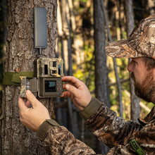 Load image into Gallery viewer, Spartan GoLive 2 in Spartan Realtree Edge. This camo has a greenish tone with leaves and tree branches. Camera is open while he setup the camera for arming.